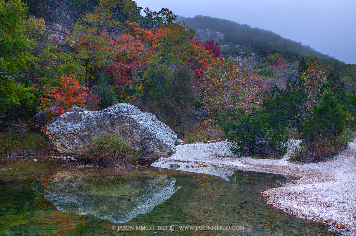 Bigtooth maple trees (Acer grandidentatum) in fall color and a boulder reflected in a pool on the Sabinal River under a layer...